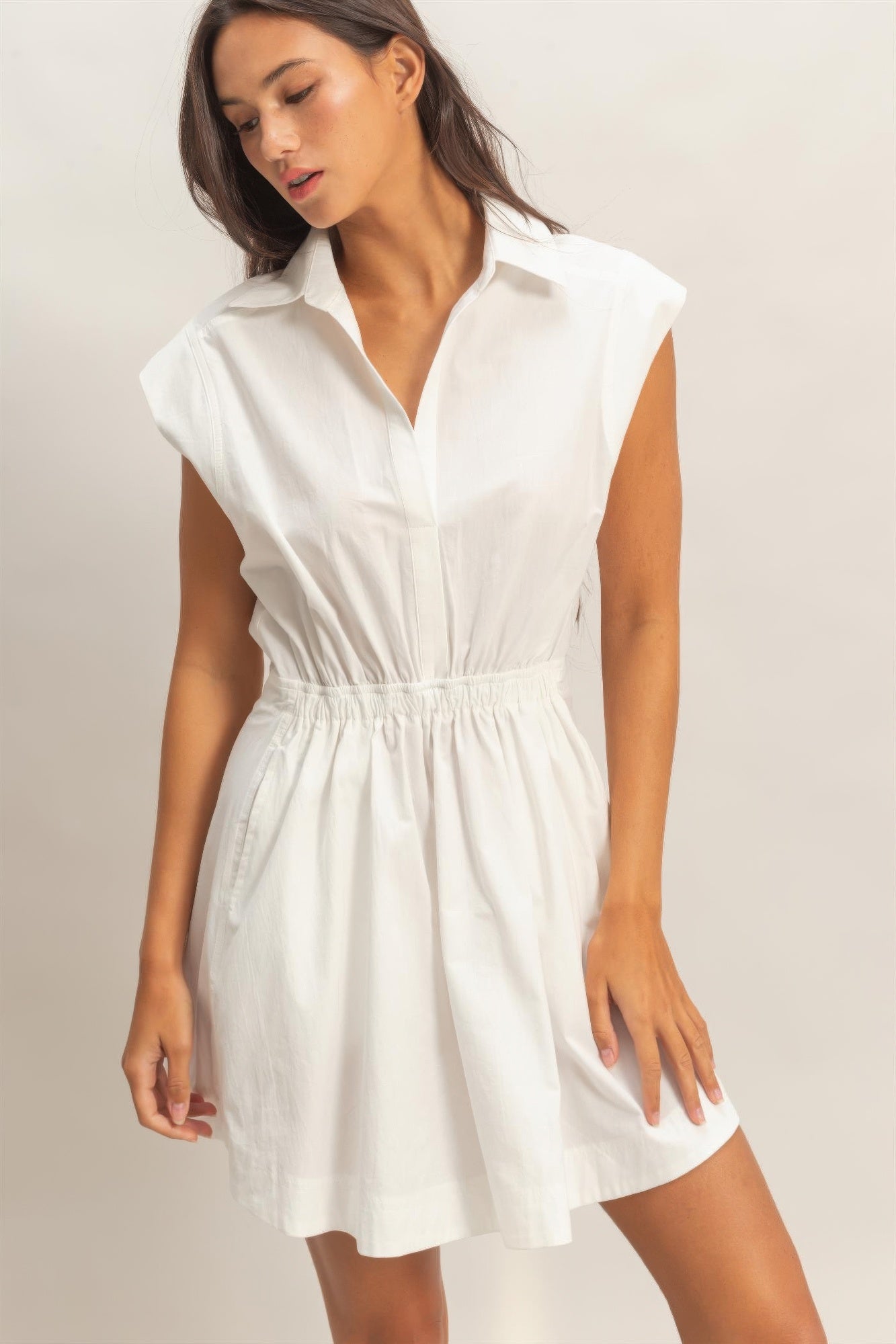 A woman posing in a white sleeveless collar mini dress with shirred waist detailing.